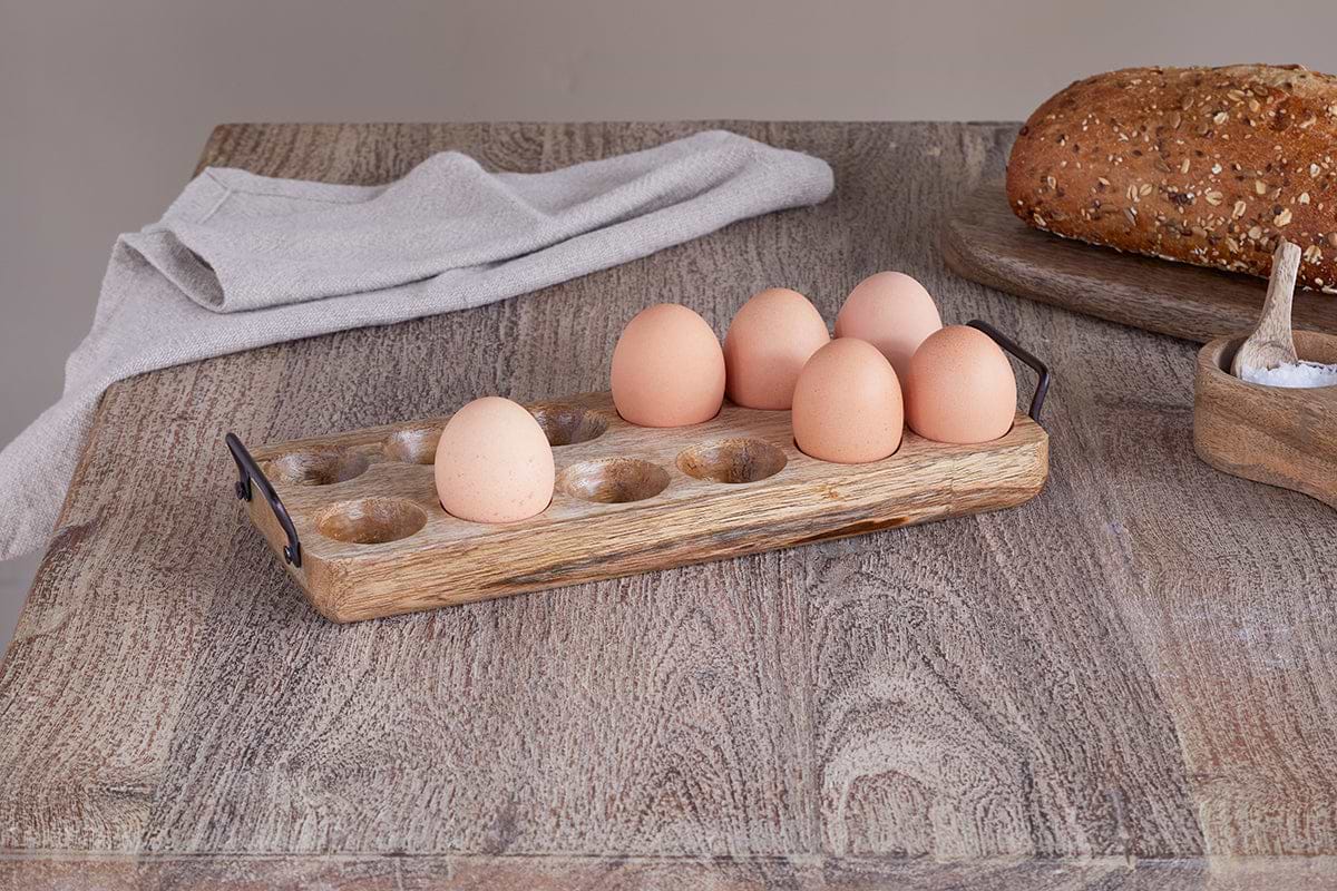 The nkuku Anda Mango Wood Egg Holder - Natural, holding six brown eggs, rests on a kitchen counter beside a seeded loaf, coarse salt in a wooden dish with spoon, and a folded beige napkin for a cozy, rustic touch.