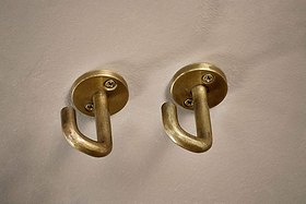 The Aniko Iron Ceiling Hook - Brass (Set of 2) by nkuku features brass-finished hooks with rounded bases and curved arms, perfect for organizing kitchen storage or hanging items, and mounted side by side on a light-colored wall.