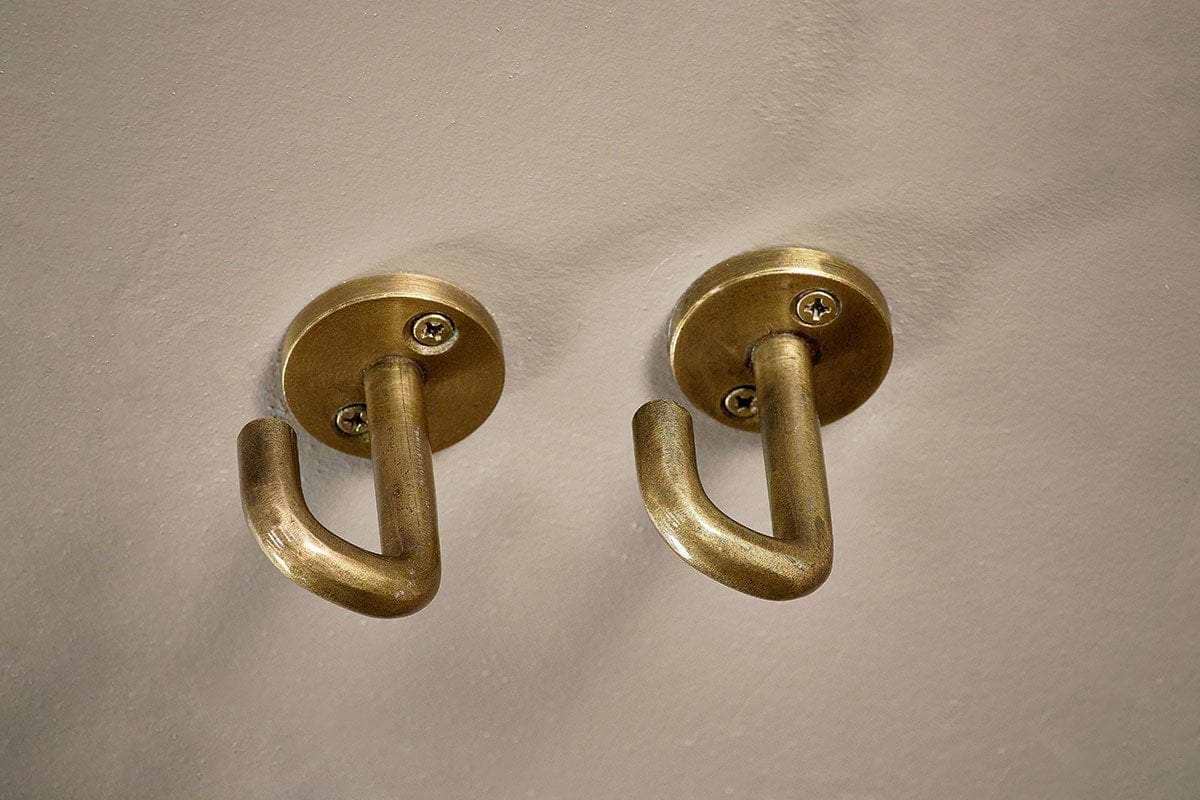 The Aniko Iron Ceiling Hook - Brass (Set of 2) by nkuku features brass-finished hooks with rounded bases and curved arms, perfect for organizing kitchen storage or hanging items, and mounted side by side on a light-colored wall.
