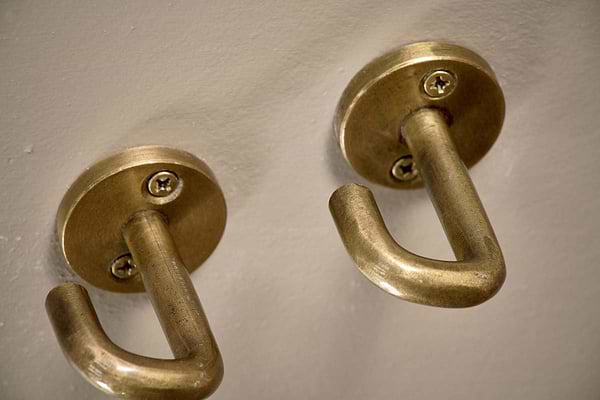 Close-up of two nkuku Aniko Iron Ceiling Hooks in brass, mounted on a light wall. Each hook curves upward for hanging, features a gently worn finish, and is secured with visible screws at the rounded base. Set of 2.