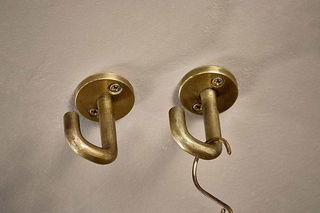 The nkuku Aniko Iron Ceiling Hook - Brass (Set of 2) is mounted side by side on a beige wall. Each hook features a round base with two visible screws. A thin metal object hangs from the right hook, while the left one remains empty.