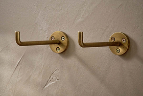 The Aniko Iron Hook - Brass (Set of 2) by nkuku features two cylindrical, right-angle brass wall hooks with three visible screws on round bases, mounted side by side for stylish hanging storage on a beige, matte-textured wall.