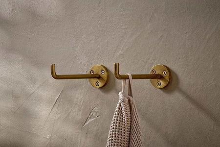 The nkuku Aniko Iron Hook - Brass (Set of 2) adds practical storage to a beige textured wall; one hook holds a woven beige towel, while the other remains empty, with light shadows and subtle imperfections enhancing the setting.