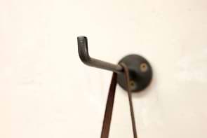 A close-up of a black, hand-cast Aniko Iron Hook by nkuku (Set of 2), mounted with two screws on a beige wall, displays a brown strap hanging from it; another L-shaped hook is slightly out of focus in the background.