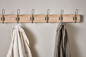 The Ankita Mango Wood & Iron Hooks rack by nkuku, featuring five antique brass hooks on natural wood, is mounted on a light wall. A white garment hangs on the leftmost hook; a gray one is second from the right. The other hooks are empty.
