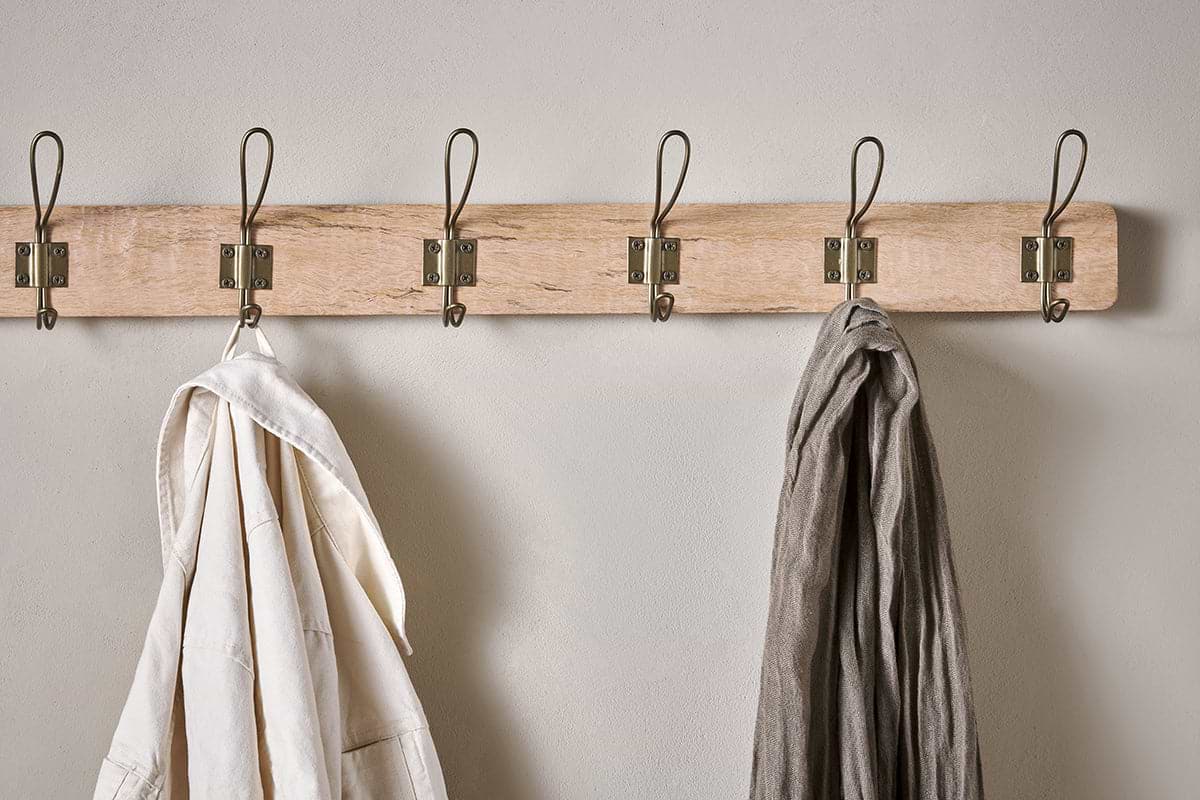 The Ankita Mango Wood & Iron Hooks rack by nkuku, featuring five antique brass hooks on natural wood, is mounted on a light wall. A white garment hangs on the leftmost hook; a gray one is second from the right. The other hooks are empty.