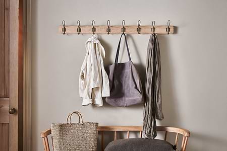 The Ankita Mango Wood & Iron Hooks by nkuku in natural and antique brass hold a cream jacket, grey tote bag, and grey scarf. Below, a woven tote rests on a wooden bench with a dark grey cushion.