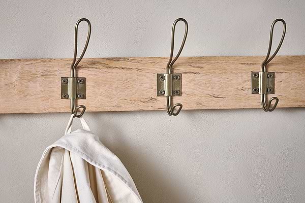 The Nkuku Ankita Mango Wood & Iron Hooks - Natural & Antique Brass - Small features three artisan metal hooks mounted on a wooden rack. The left hook holds a cream-colored cloth or bag, while the other two are empty and attached with visible screws.