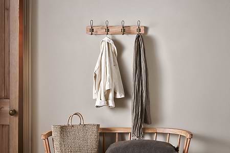The nkuku Ankita Mango Wood & Iron Hooks in Natural & Antique Brass (Small) are mounted above a wooden bench. A light shirt and gray scarf hang from the hooks, with a woven bag and dark cushion nearby against a light gray wall.