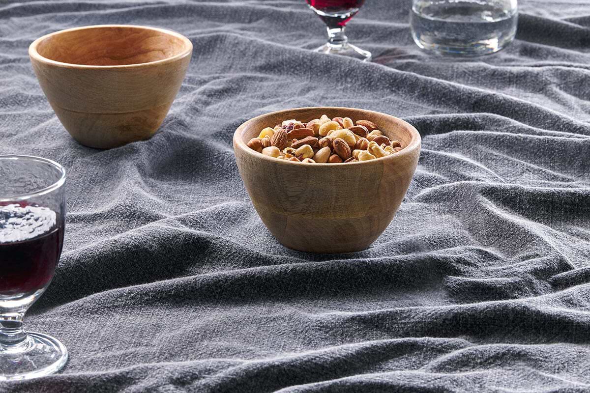 The nkuku Artisan Bowl, filled with mixed nuts, rests on textured gray fabric beside an empty bowl, a glass of red wine, and water—creating an inviting tabletop setting with stylish accessories.