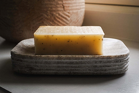 A yellow soap bar with dark flecks rests on the nkuku Asana Marble Soap Dish - Grey, displayed on a light surface in gentle natural light.