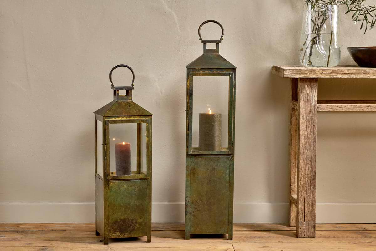 Two tall, rustic Asharu Lanterns - Antique Patina by nkuku, featuring glass panels and an aged finish, sit on a wooden floor and make a striking statement piece. Each lantern holds a lit pillar candle inside. The larger lantern is positioned on the right. A wooden table with a glass vase and plant is partially visible to the right.