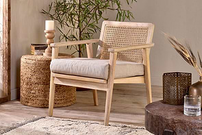 The nkuku Atri Mango Wood & Cane Occasional Chair, with a woven rattan backrest and beige cushion, rests on a light rug beside a round wicker side table holding a candle and vase, accented by plants for a cozy, natural look.