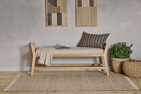 The Avanthi Upholstered Sleigh Bench by nkuku, with natural linen upholstery, sits on a jute rug. Above, woven wall art panels hang. A terracotta potted plant and wicker basket complete the boho-neutral look with chevron pillow and throw.