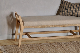 The nkuku Avanthi Upholstered Sleigh Bench in natural mango wood features a beige cushion and sits on a wooden floor, paired with a textured woven rug and a black and gold chevron pillow against a light gray wall.