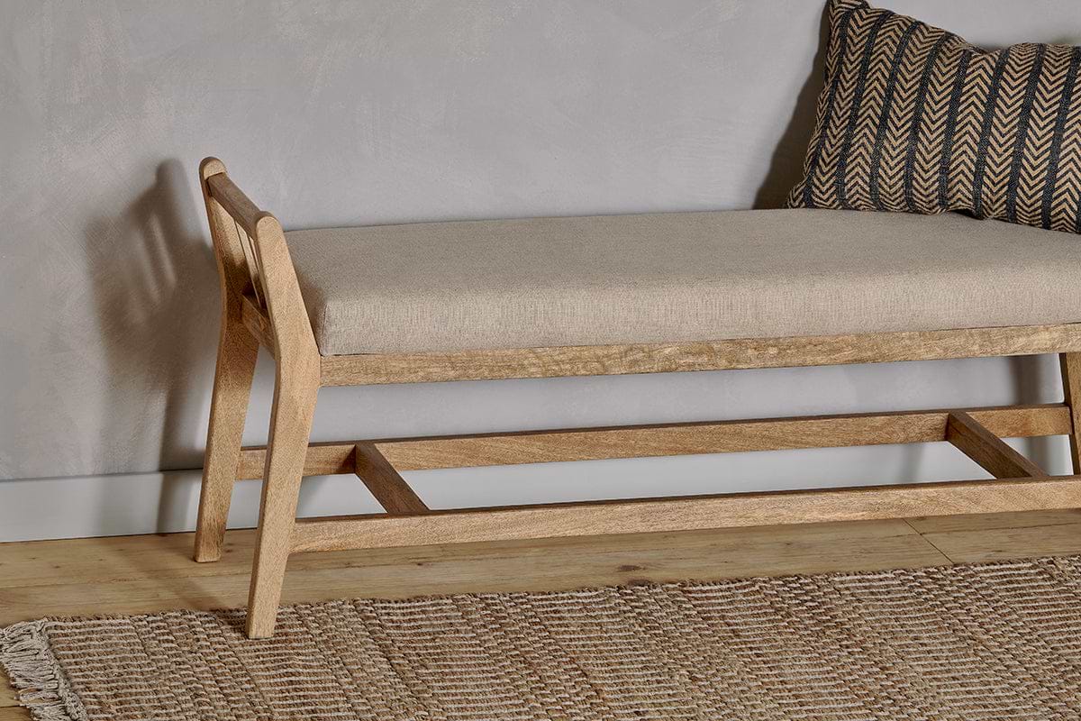 The nkuku Avanthi Upholstered Sleigh Bench in natural mango wood features a beige cushion and sits on a wooden floor, paired with a textured woven rug and a black and gold chevron pillow against a light gray wall.