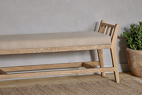 The Avanthi Upholstered Sleigh Bench by nkuku, crafted from hand-carved mango wood with a slatted backrest and natural linen seat, sits on a woven rug beside a textured gray wall.