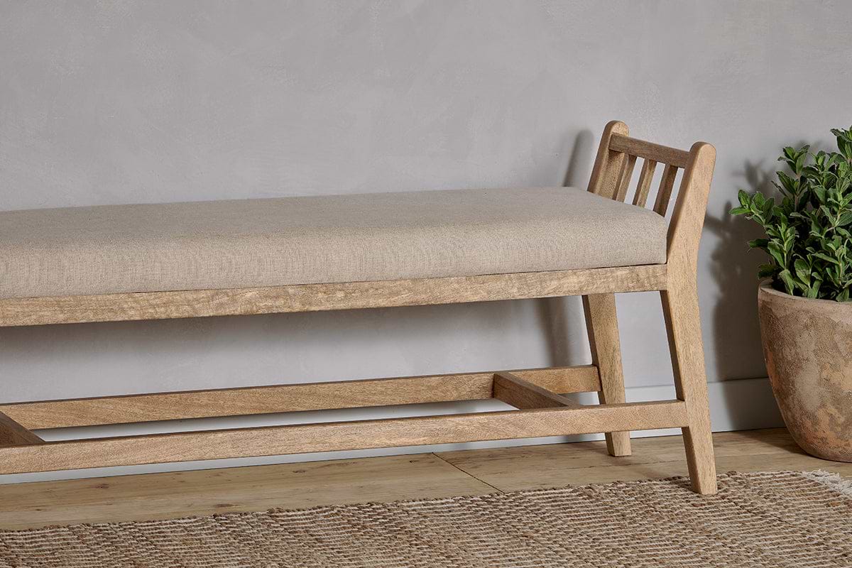 The Avanthi Upholstered Sleigh Bench by nkuku, crafted from hand-carved mango wood with a slatted backrest and natural linen seat, sits on a woven rug beside a textured gray wall.