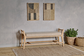 The nkuku Avanthi Upholstered Sleigh Bench - Natural, featuring a beige cushion, rests by a light gray wall. Above it are two abstract textile art pieces; beside the bench on the woven rug and light wood floor sit a green plant and woven basket.