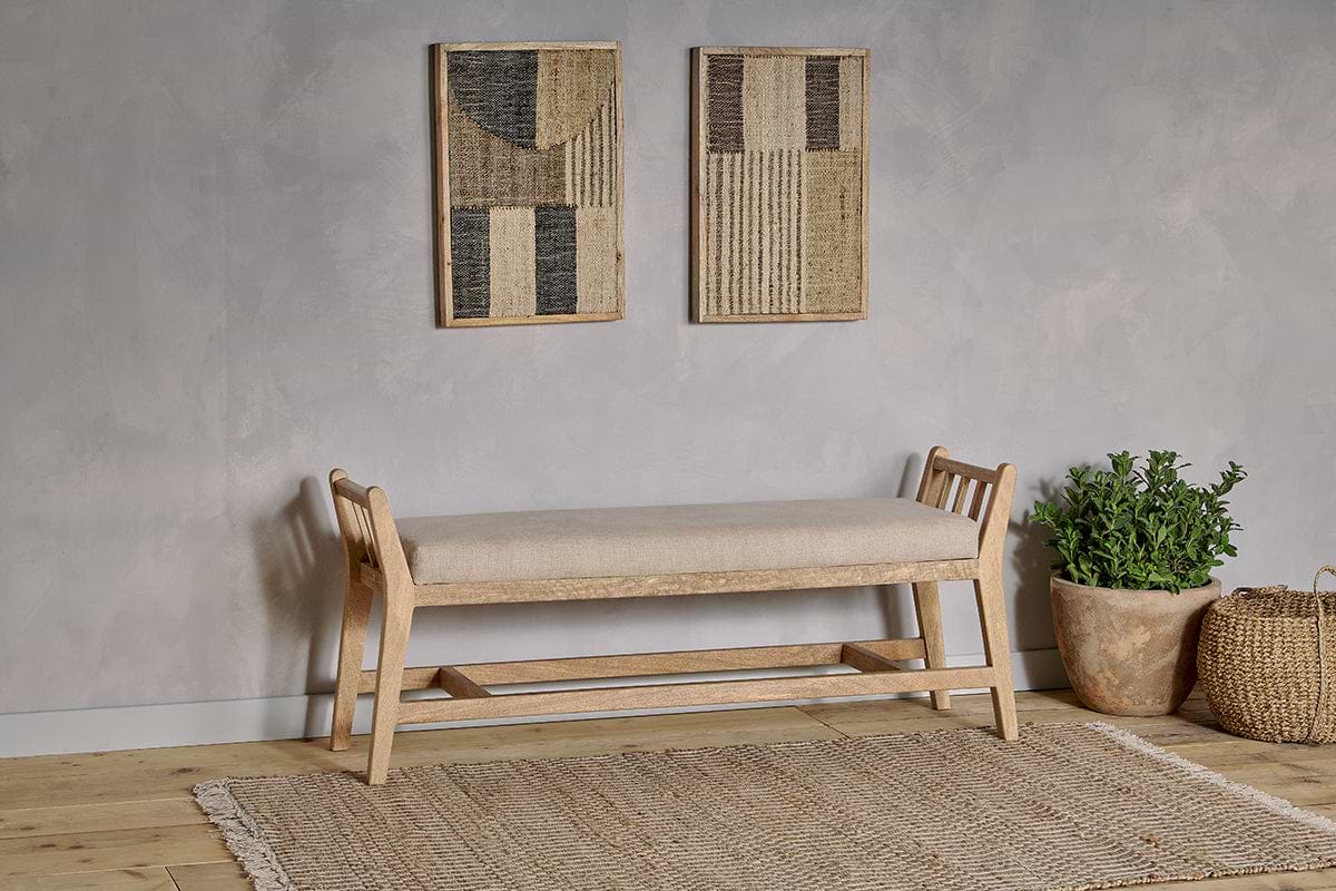 The nkuku Avanthi Upholstered Sleigh Bench - Natural, featuring a beige cushion, rests by a light gray wall. Above it are two abstract textile art pieces; beside the bench on the woven rug and light wood floor sit a green plant and woven basket.