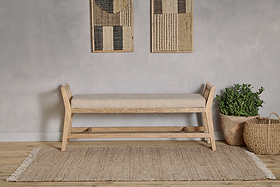The Avanthi Upholstered Sleigh Bench by nkuku in natural linen rests on a woven rug, complemented by a gray textured wall, abstract art, a green potted plant in a clay pot, and a woven basket for modern neutral decor.