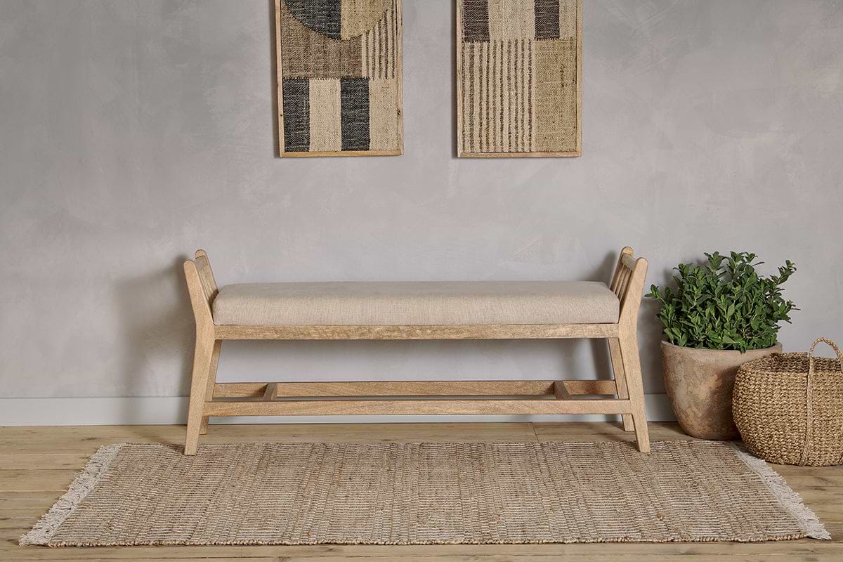 The Avanthi Upholstered Sleigh Bench by nkuku in natural linen rests on a woven rug, complemented by a gray textured wall, abstract art, a green potted plant in a clay pot, and a woven basket for modern neutral decor.