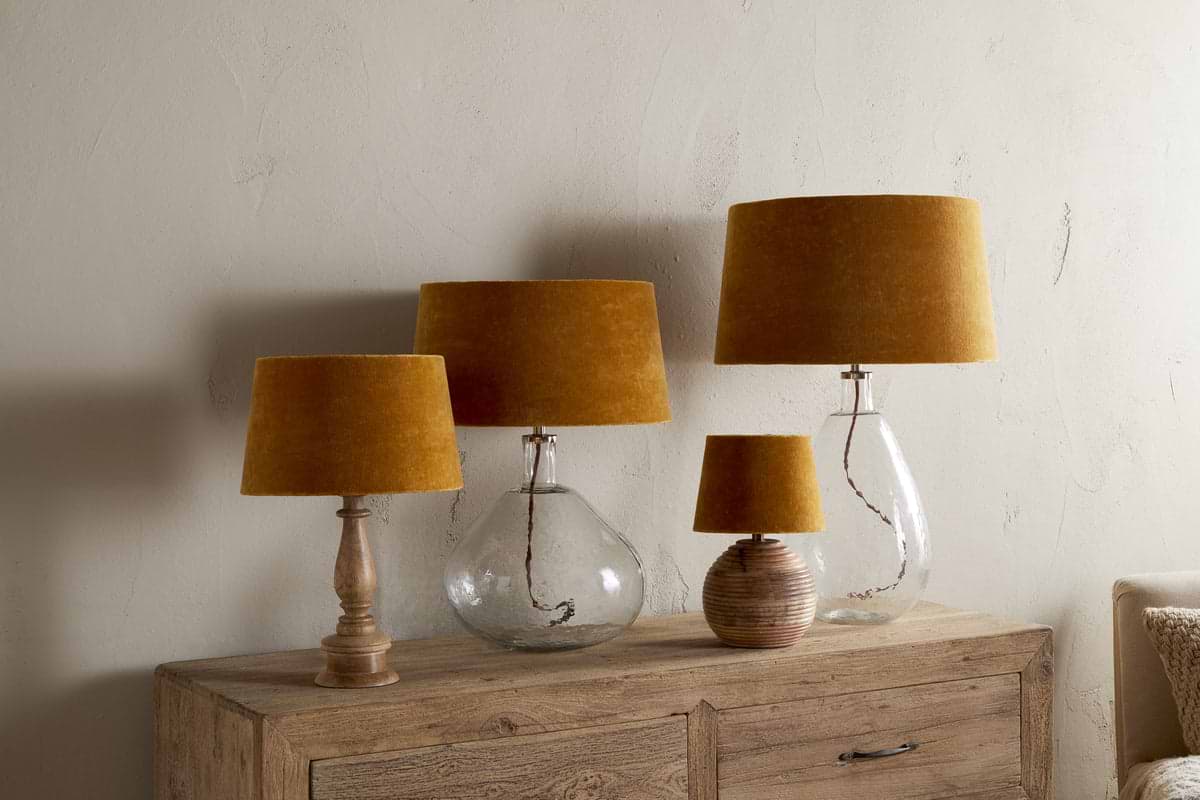 Four table lamps with Batakha Velvet & Linen Lampshades in Dark Ochre by nkuku are arranged on a wooden sideboard; two have clear glass bases, one has a ribbed ceramic base, and one features a turned wooden base, set against a beige wall.