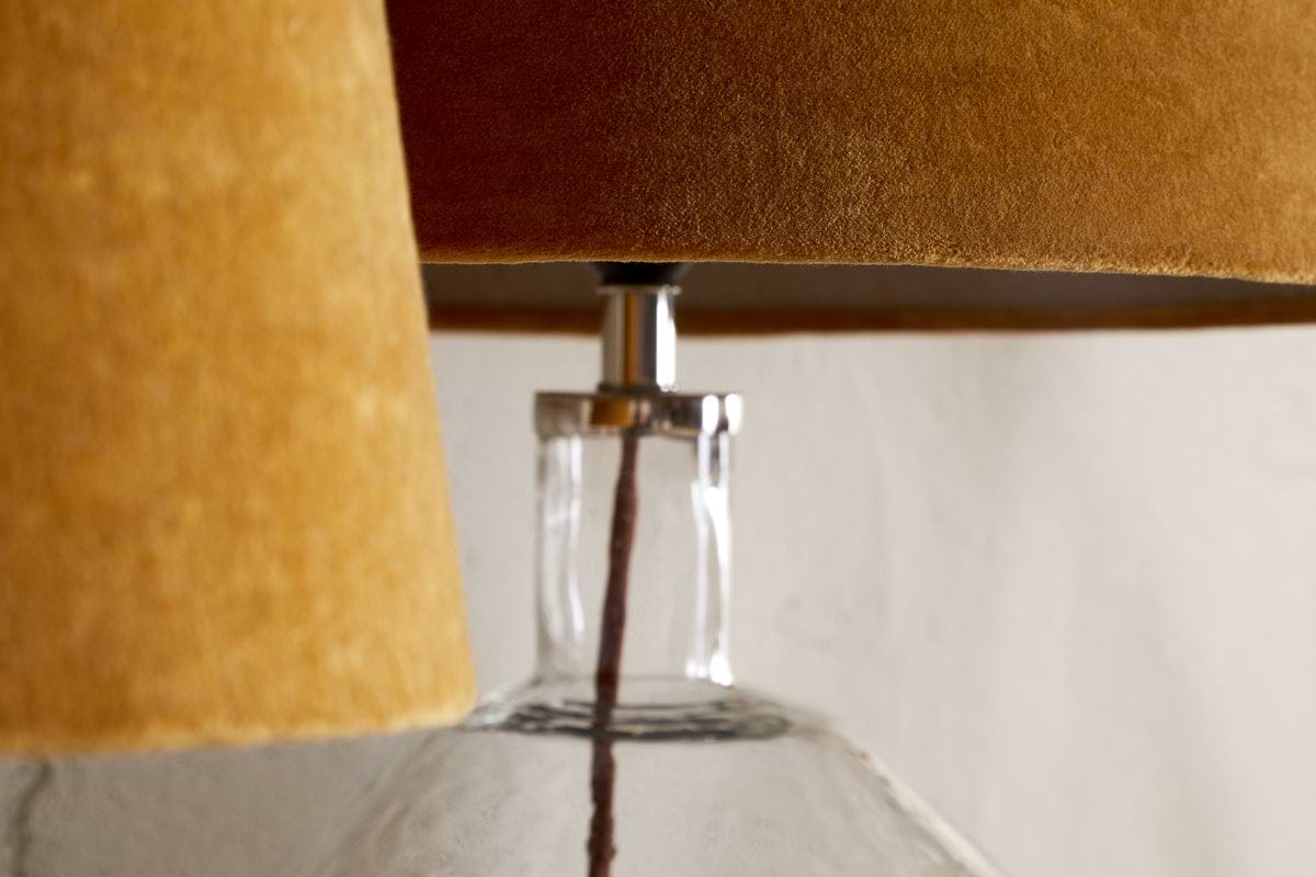 Close-up of two overlapping nkuku Batakha Velvet & Linen Lampshades in Dark Ochre on a clear glass lamp base, highlighting their texture and detail against a neutral background.
