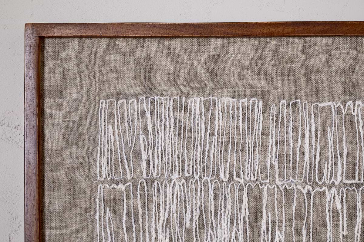 The Birala Hand Stitched Wall Art - Natural by nkuku displays irregular vertical white threads on beige fabric, framed in sustainable mango wood, highlighting the textured abstract pattern against a softly textured wall.