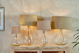 Four nkuku Baba Recycled Glass Lamps in Clear, Large Wide, with beige fabric shades, are arranged on a wooden console. Their recycled glass bases reveal twisted brown cords, casting a soft, warm glow against a textured beige wall.