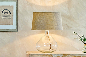 The Baba Recycled Glass Lamp - Clear - Large Wide by nkuku, featuring an elegant base and beige fabric shade, sits on a light wooden console with textured neutral walls, abstract art, and a small vase with green foliage.
