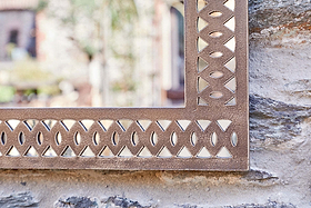 A close-up of the Bahula Decorative Outdoor Mirror - Rust by nkuku shows its metallic geometric frame reflecting light. Mounted on textured stone outdoors, it softly mirrors a blurred garden scene in the background.