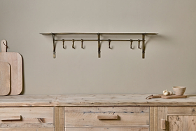 A rustic wood counter displays cutting boards, a garlic bulb, and a wooden bowl. Above, nkuku's Basti Iron Storage Shelf in Antique Brass with movable hooks is mounted on the beige wall for a minimalist look.