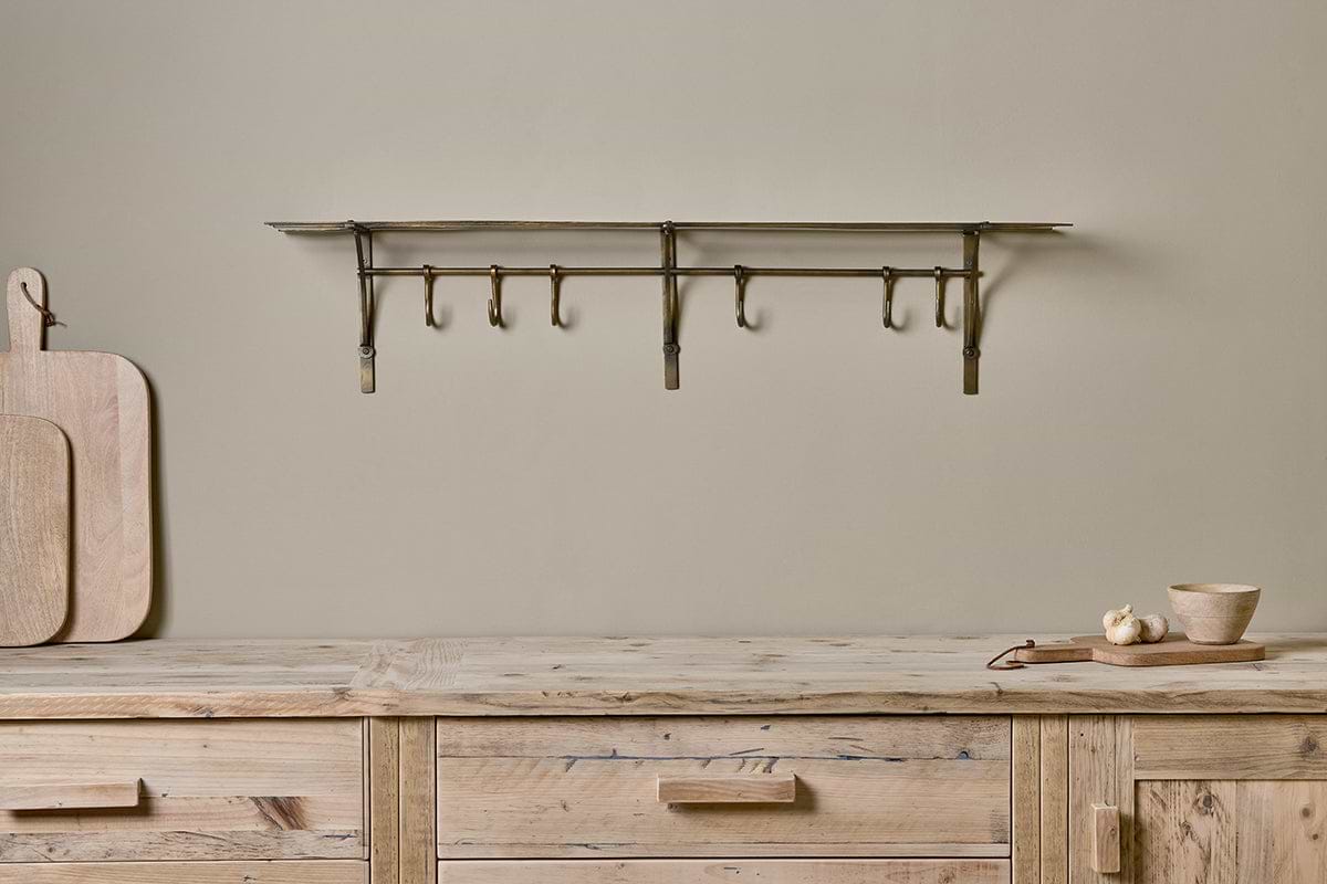 A rustic wood counter displays cutting boards, a garlic bulb, and a wooden bowl. Above, nkuku's Basti Iron Storage Shelf in Antique Brass with movable hooks is mounted on the beige wall for a minimalist look.