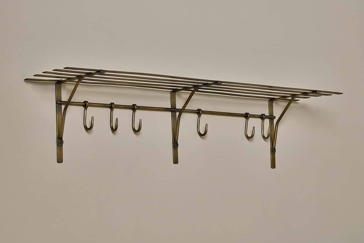 The nkuku Basti Iron Storage Shelf in antique brass features a vintage-style slatted top shelf and six hooks, mounted on a light wall for classic elegance and sturdy storage.