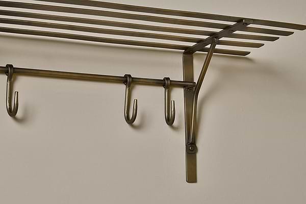 A close-up of the nkuku Basti Iron Storage Shelf in antique brass, mounted on a beige wall, features horizontal slats as a shelf and a bar below with three S-hooks for hanging. Visible screws and brackets provide sturdy support.