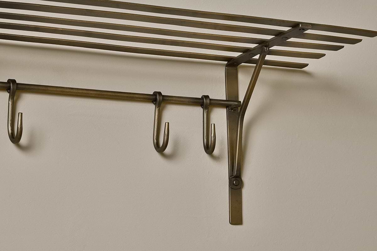 A close-up of the nkuku Basti Iron Storage Shelf in antique brass, mounted on a beige wall, features horizontal slats as a shelf and a bar below with three S-hooks for hanging. Visible screws and brackets provide sturdy support.