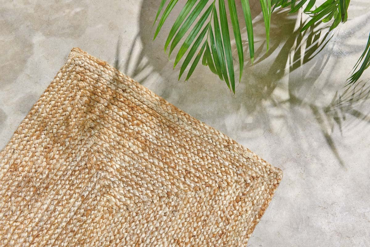 A close-up of the nkuku Braided Hemp Floor Runner displays its woven texture and braided pattern on a light concrete floor, while green palm leaves in the corner cast soft shadows, enhancing the natural, relaxed vibe.