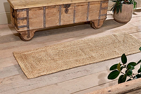 The nkuku Braided Hemp Floor Runner, a rectangular eco-friendly piece, lies on a light wooden floor by a rustic chest. Green potted plants at the edges enhance the cozy, natural look.
