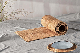 A rolled-up nkuku Braided Hemp Table Runner is unrolled on a light gray textured surface, with a blue-gray ceramic plate on a matching round placemat nearby, creating an artisanal tablescape accented by dried grasses.