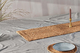 A light gray cloth-covered table features artisan tableware, a brass candle holder with a mauve candle, and the nkuku Braided Hemp Table Runner with a matching placemat beneath a blue-gray ceramic plate. Dried grass accents the tablescape.