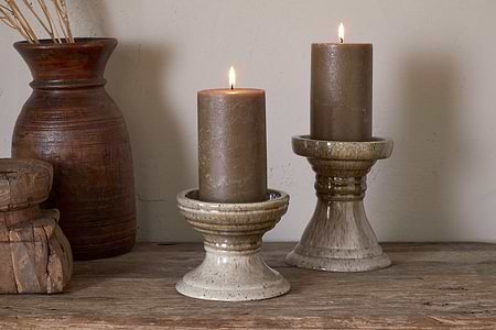 Two brown candles glow on nkuku’s Cabra Reactive Glaze Candle Holders in Olive & Natural, each at a different height. Set atop rustic wood with a wooden vase of dried grasses, they bring cozy, earthy style to a neutral wall.