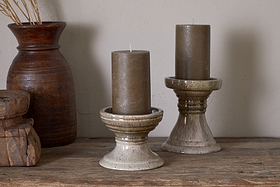 Two brown pillar candles sit in nkuku Cabra Reactive Glaze Candle Holders in Olive & Natural on a wooden surface, with a tall brown clay vase of dried grass and stacked wooden boxes creating an earthy, rustic vintage decor.