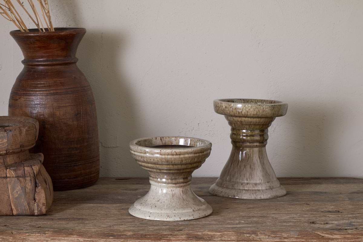 Two nkuku Cabra Reactive Glaze Candle Holders in olive and natural rest on wood. A tall, dark brown vase with dried grasses and a rustic wooden box add to the decor against a light beige textured wall.