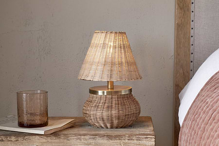 The nkuku Cija Rattan Table Lamp - Natural, featuring a woven rattan shade and brass detail, sits on a rustic nightstand beside a brown glass tumbler and closed book, next to a bed with blush bedding.