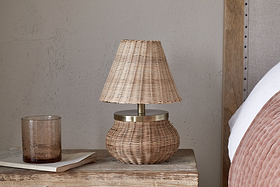 The nkuku Cija Rattan Table Lamp - Natural, with a woven shade and brass accent, sits on a rustic nightstand beside a brown glass tumbler, a closed book, and a bed with a pink pillow.
