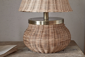 The Cija Rattan Table Lamp - Natural by nkuku, featuring a wide rounded base and matching rattan shade with brass accents, rests on a wooden surface beside a closed book.