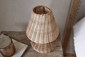 The Cija Rattan Table Lamp by nkuku, with its tiered woven shade, rests on a rustic wooden bedside table beside a brown glass container and a closed book.