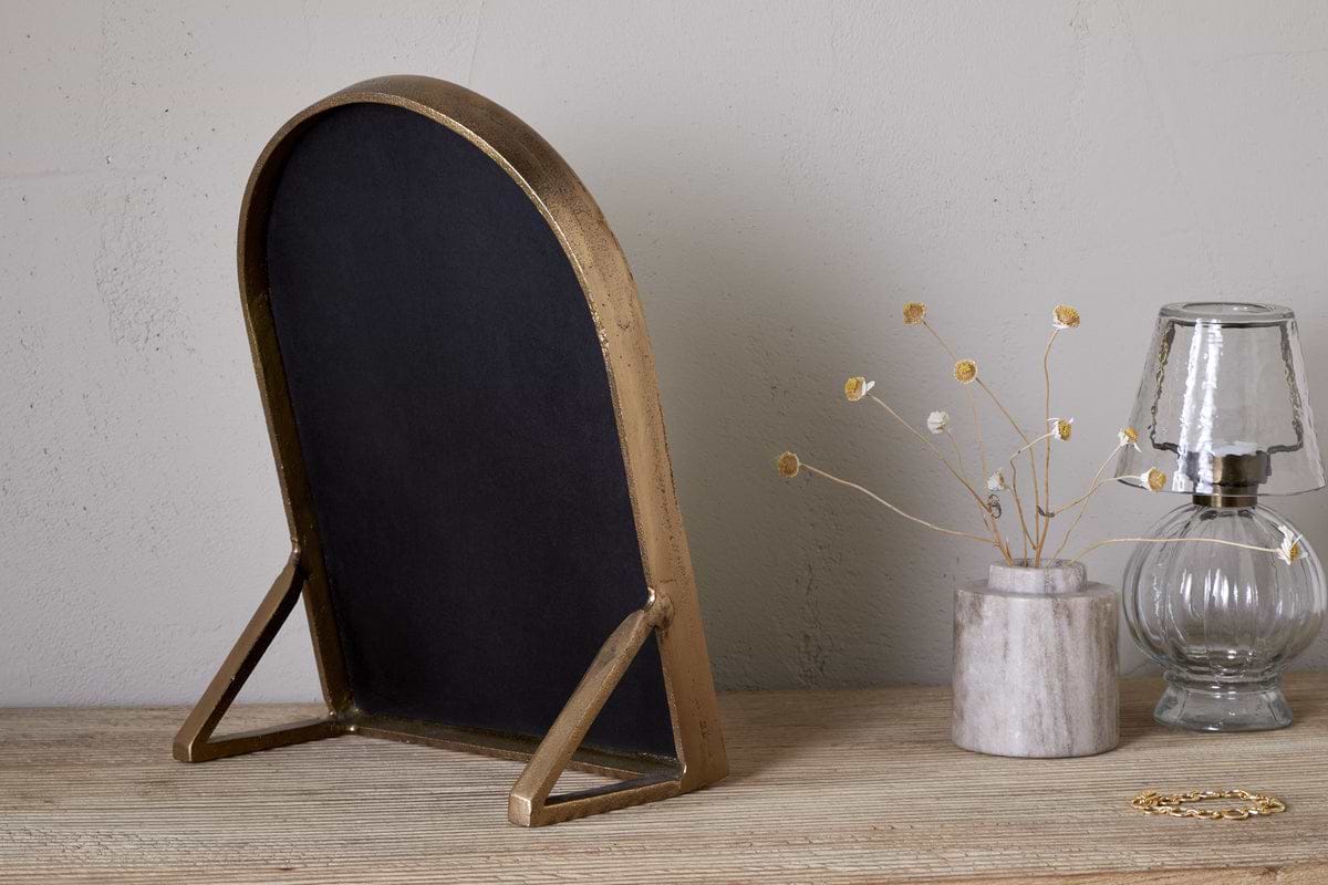 The nkuku Cayce Cast Aluminum Vanity Mirror in Antique Brass stands on a wooden dressing table beside a marble vase with dried flowers, a clear glass oil lamp, and a gold bracelet. The background features a neutral, plain wall.
