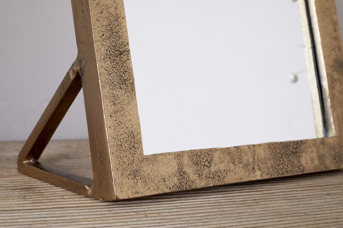 Close-up of the nkuku Cayce Cast Aluminum Vanity Mirror in Antique Brass, featuring a rectangular, textured frame resting on a light wooden surface. Ideal as a dressing table or standing vanity mirror.
